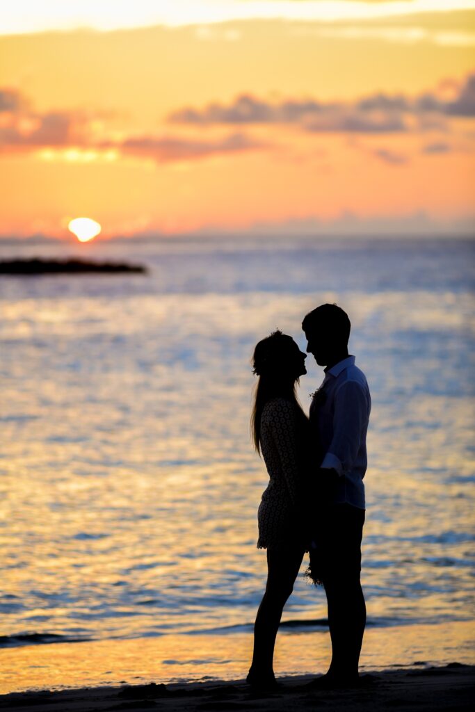 Image of couple standing on beachfront as the sun sets over the water. 